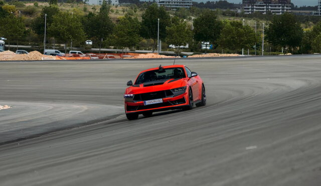 Red sports car drifting around a race track, tire marks visible on the asphalt with green trees in the background.