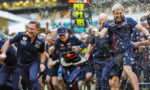 Red Bull racing team members sprint at a race start, water and confetti flying around as a scoreboard glows in the background.
