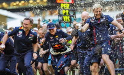 Red Bull racing team members sprint at a race start, water and confetti flying around as a scoreboard glows in the background.