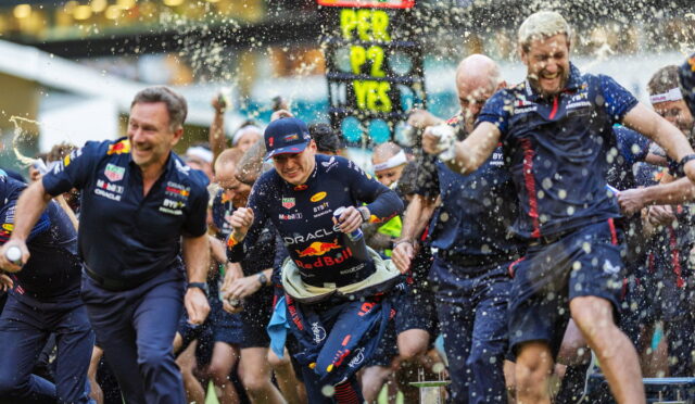 Red Bull racing team members sprint at a race start, water and confetti flying around as a scoreboard glows in the background.
