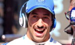 Smiling man wearing a blue cap and white racing suit with over-ear headphones, likely in a pit area or broadcasting setup.