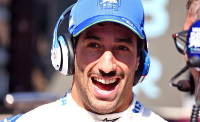 Smiling man wearing a blue cap and white racing suit with over-ear headphones, likely in a pit area or broadcasting setup.