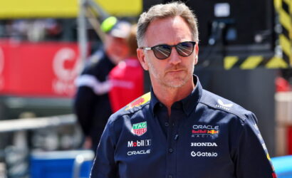 Man in dark blue racing team shirt with sponsor logos and sunglasses at a motorsport pit area, looking off to the side with a serious expression.