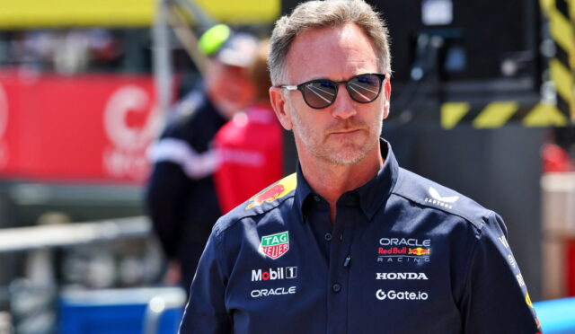 Man in dark blue racing team shirt with sponsor logos and sunglasses at a motorsport pit area, looking off to the side with a serious expression.