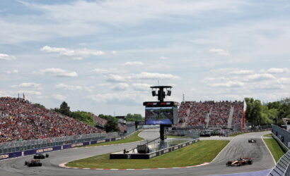 Wide view of a Formula 1 race track with cars navigating a curve, crowds in the grandstands, and a large scoreboard in the center