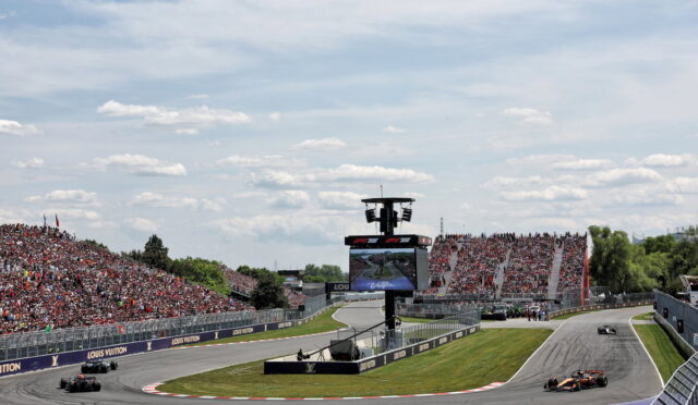Wide view of a Formula 1 race track with cars navigating a curve, crowds in the grandstands, and a large scoreboard in the center