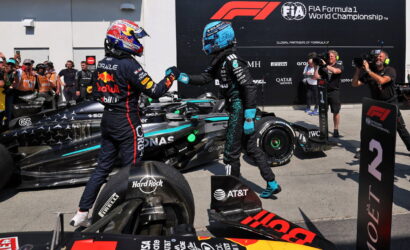 Two Formula 1 drivers share a fist bump beside their race cars on the pit lane, with photographers and a FIA banner in the background.