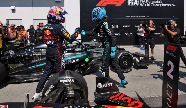 Two Formula 1 drivers share a fist bump beside their race cars on the pit lane, with photographers and a FIA banner in the background.