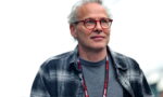 Man with gray hair and red glasses, wearing a gray shirt and plaid jacket, looking slightly upward at an outdoor event.