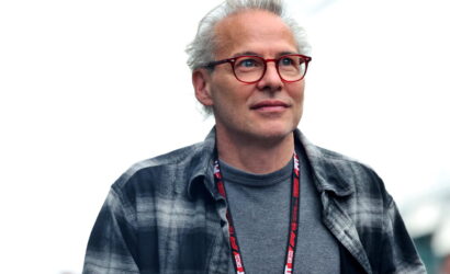 Man with gray hair and red glasses, wearing a gray shirt and plaid jacket, looking slightly upward at an outdoor event.