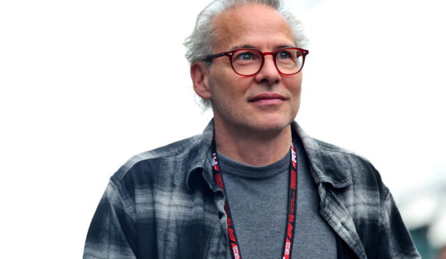 Man with gray hair and red glasses, wearing a gray shirt and plaid jacket, looking slightly upward at an outdoor event.