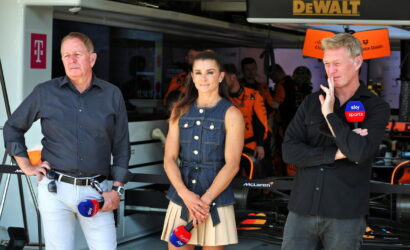 Three presenters stand in a race garage; a left man in a dark shirt, center woman in a denim vest and beige skirt, right man with Sky Sports mic.