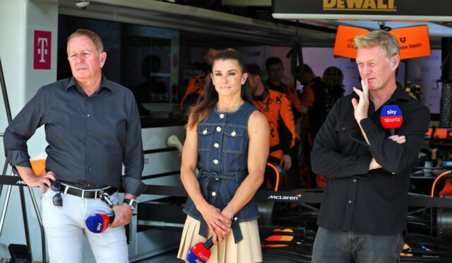 Three presenters stand in a race garage; a left man in a dark shirt, center woman in a denim vest and beige skirt, right man with Sky Sports mic.