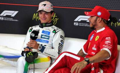 Two Formula 1 drivers sit on a couch during an interview; one in white racing suit holding a microphone, the other in red, chatting off to the side.
