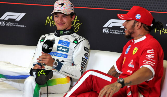 Two Formula 1 drivers sit on a couch during an interview; one in white racing suit holding a microphone, the other in red, chatting off to the side.