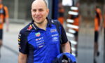 Smiling man in a blue Red Bull Racing polo with sponsor logos, outdoors in an urban area, carrying a backpack and a blue helmet bag.