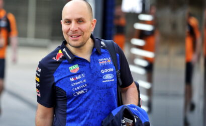 Smiling man in a blue Red Bull Racing polo with sponsor logos, outdoors in an urban area, carrying a backpack and a blue helmet bag.