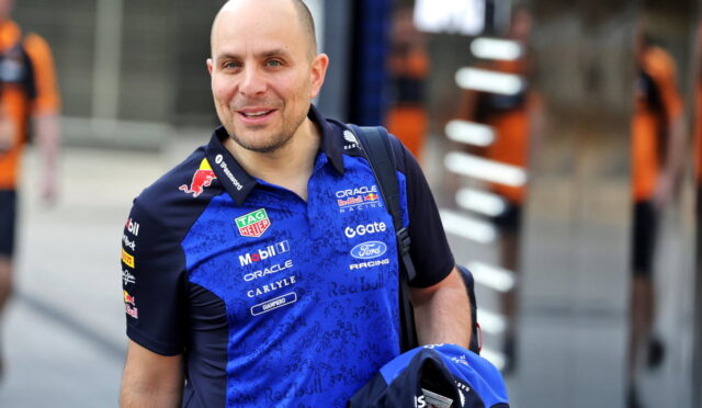 Smiling man in a blue Red Bull Racing polo with sponsor logos, outdoors in an urban area, carrying a backpack and a blue helmet bag.