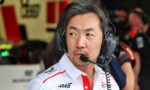Race team engineer wearing a headset with microphone in a pit garage, looking to the side in a white shirt with sponsor logos.