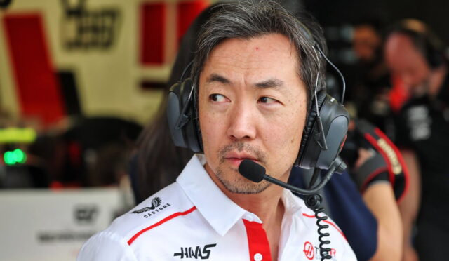 Race team engineer wearing a headset with microphone in a pit garage, looking to the side in a white shirt with sponsor logos.