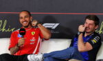Two men in racing team uniforms sit on a white couch during a press event; the left man in a red Ferrari-style shirt holds a mic and smiles, the right man in blue laughs.