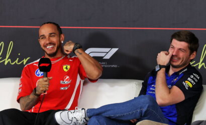 Two men in racing team uniforms sit on a white couch during a press event; the left man in a red Ferrari-style shirt holds a mic and smiles, the right man in blue laughs.