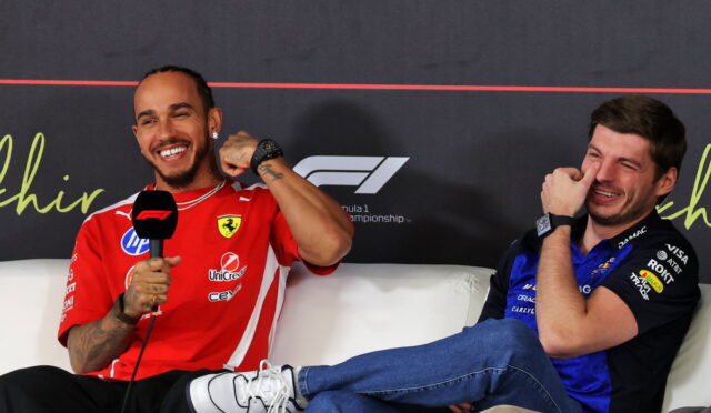 Two men in racing team uniforms sit on a white couch during a press event; the left man in a red Ferrari-style shirt holds a mic and smiles, the right man in blue laughs.