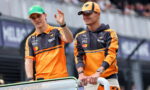 Two McLaren team members in orange uniforms wave from a race car on the track with fans in the background.