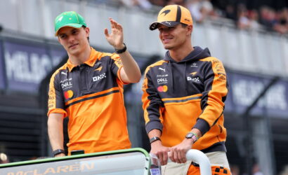 Two McLaren team members in orange uniforms wave from a race car on the track with fans in the background.