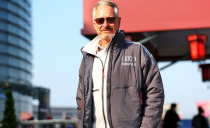 Man with gray hair and sunglasses in a gray zip-up jacket with Audi Revolut logo, standing outdoors in an urban area.
