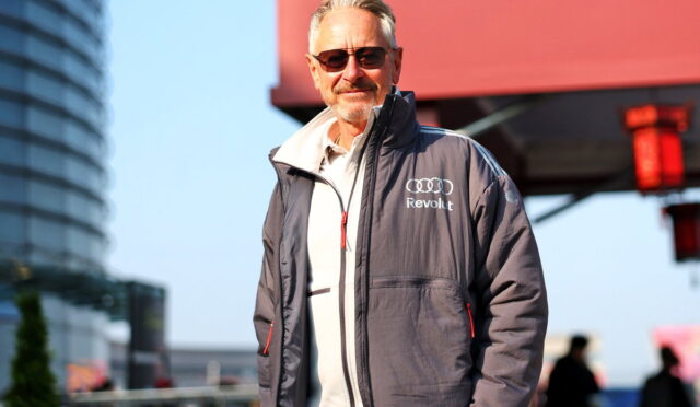 Man with gray hair and sunglasses in a gray zip-up jacket with Audi Revolut logo, standing outdoors in an urban area.