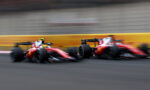 Two red Formula 1 cars race side by side on a track, blurred by motion with a yellow barrier in the background.