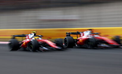 Two red Formula 1 cars race side by side on a track, blurred by motion with a yellow barrier in the background.