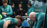 Team members in teal uniforms surround a race car in the Aston Martin garage, discussing strategy.