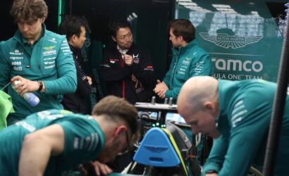 Team members in teal uniforms surround a race car in the Aston Martin garage, discussing strategy.