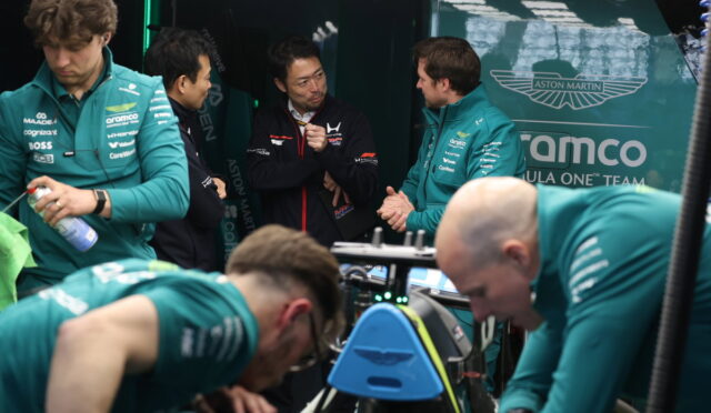 Team members in teal uniforms surround a race car in the Aston Martin garage, discussing strategy.