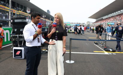 Two TV presenters interview each other in a race track pit lane, microphones in hand, with equipment and spectators in the background.