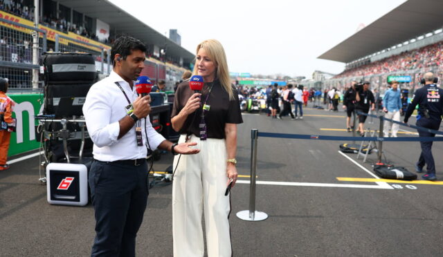 Two TV presenters interview each other in a race track pit lane, microphones in hand, with equipment and spectators in the background.