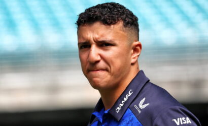 Close-up portrait of a man with short curly hair in a blue team polo, a serious expression, with stadium seating in the blurred background.