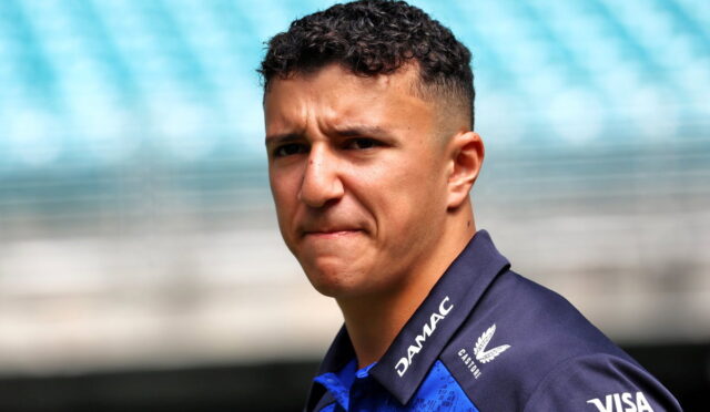 Close-up portrait of a man with short curly hair in a blue team polo, a serious expression, with stadium seating in the blurred background.