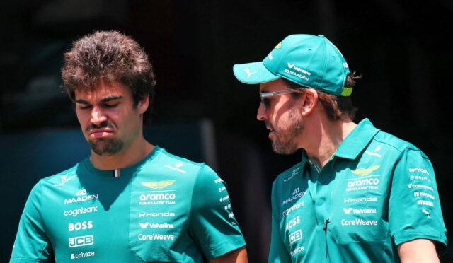 Two men in teal team uniforms stand outdoors; one on the left squints with a frown while the other on the right wears a teal cap and speaks to him.