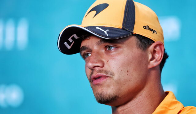 Close-up of a man wearing a yellow cap and orange shirt, looking off to the side against a blue background.