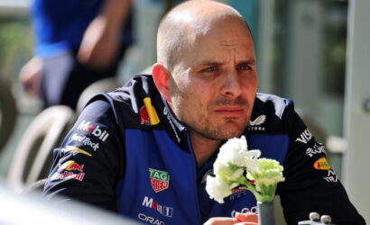 Man in a blue and black racing suit sits at a table outdoors, looking pensive, with a small vase of white flowers in front.