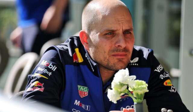 Man in a blue and black racing suit sits at a table outdoors, looking pensive, with a small vase of white flowers in front.
