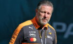 Man with gray hair and beard wearing an orange and black racing team polo with sponsor logos, looking off to the side in daylight.