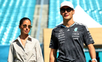 Two people walk in a sunny stadium; a woman on the left wears a beige checkered shirt and sunglasses, while a man on the right wears a black racing jersey and a white cap.