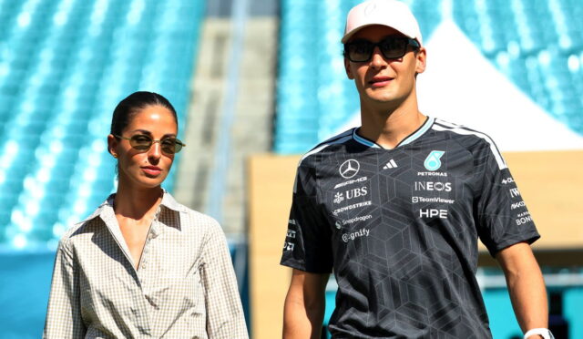 Two people walk in a sunny stadium; a woman on the left wears a beige checkered shirt and sunglasses, while a man on the right wears a black racing jersey and a white cap.
