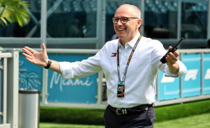 Smiling man in a white shirt with a lanyard and walkie-talkie, arms raised in a welcoming gesture outdoors at an event.
