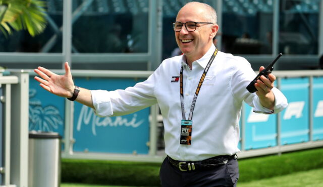 Smiling man in a white shirt with a lanyard and walkie-talkie, arms raised in a welcoming gesture outdoors at an event.