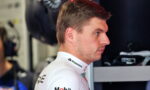 Side profile of a race car driver in a white sponsor-branded suit in a garage/pit area